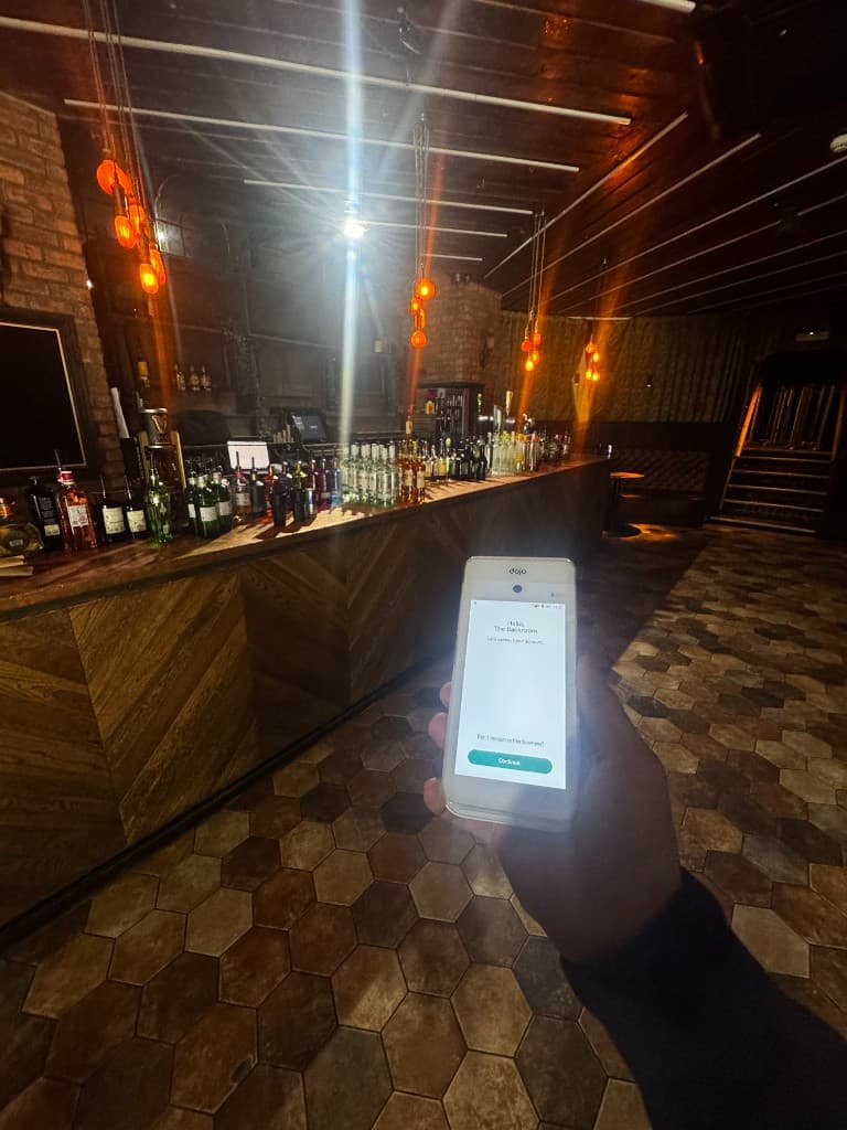 Dojo terminal held up in a dimly lit cocktail bar at one of our hospitality clients, bottles lined along the bar