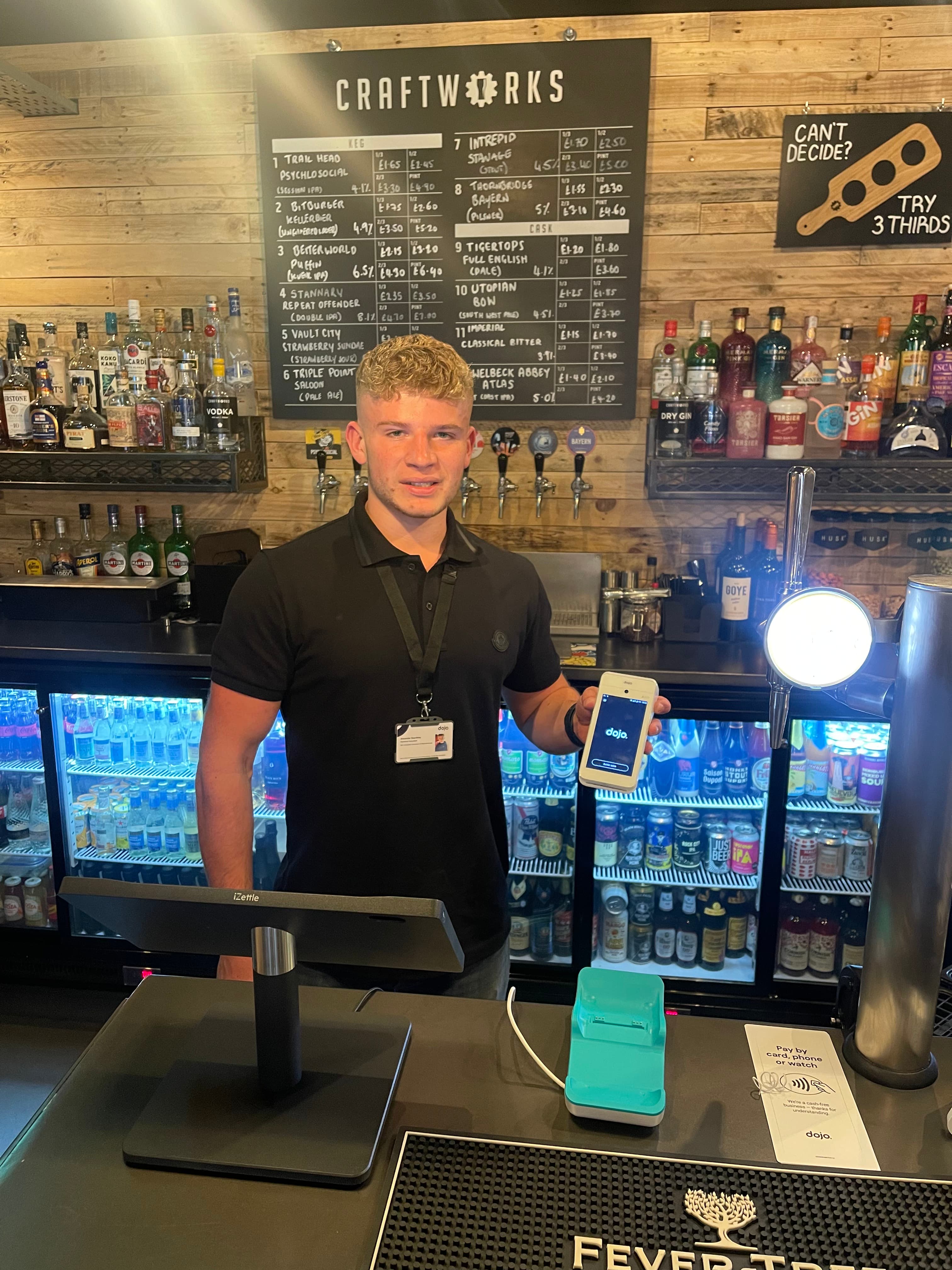 Taproom staff member holding a Dojo terminal in front of a chalkboard beer menu at one of our brewery clients